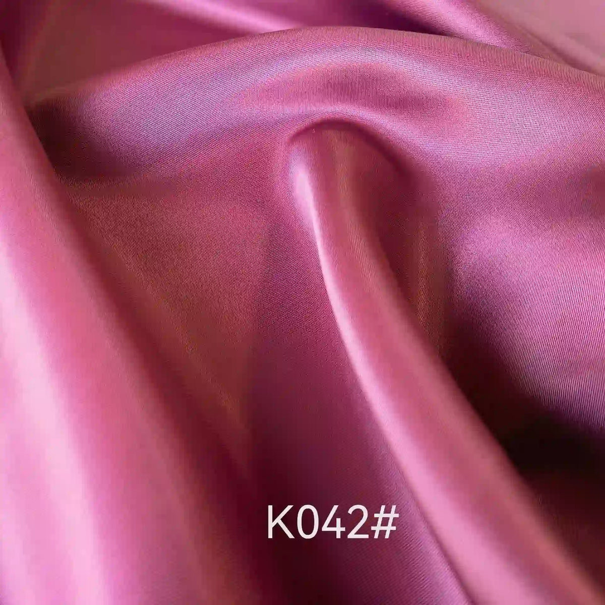 Un gros plan du tissu satiné rose lisse et brillant coloré en viscose au mètre de Longan Craft France montre des plis élégants. La mention "K042#" apparaît en bas au centre. Ce tissu est idéal pour les robes de cérémonie et est fabriqué à partir d'un tissage sergé en polyester-viscose.