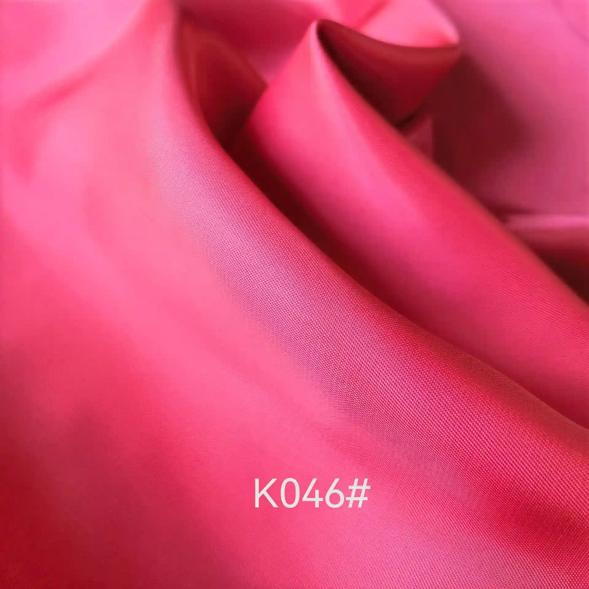 Gros plan sur le Tissu satiné coloré en viscose au mètre, rose, lisse et brillant, de Longan Craft France, présentant des plis souples et le code blanc "K046#" en bas au centre. Idéal pour les robes de cérémonie élégantes avec un tissage sergé polyester-viscose.