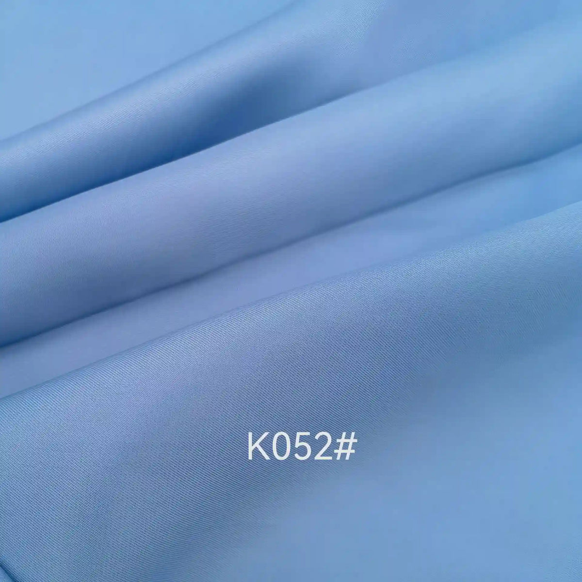 Gros plan sur le Tissu satiné coloré en viscose au mètre de Longan Craft France, de couleur bleu clair, aux plis subtils et à la texture lisse. La mention "K052#" est imprimée en blanc sur l'élégant tissu sergé en polyester-viscose, idéal pour les robes de cérémonie.