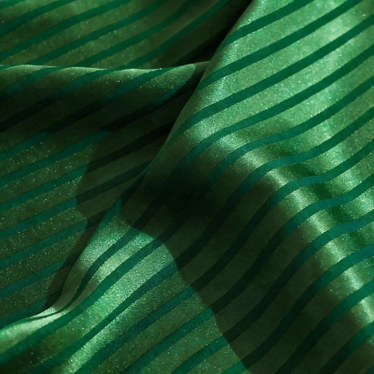 Un gros plan du tissu satin jacquard rayé coloré au mètre de Longan Craft France montre un satin lisse et brillant avec des rayures diagonales vert foncé et des plis souples créant des ombres et des lumières subtiles - idéal pour la confection de robes.
