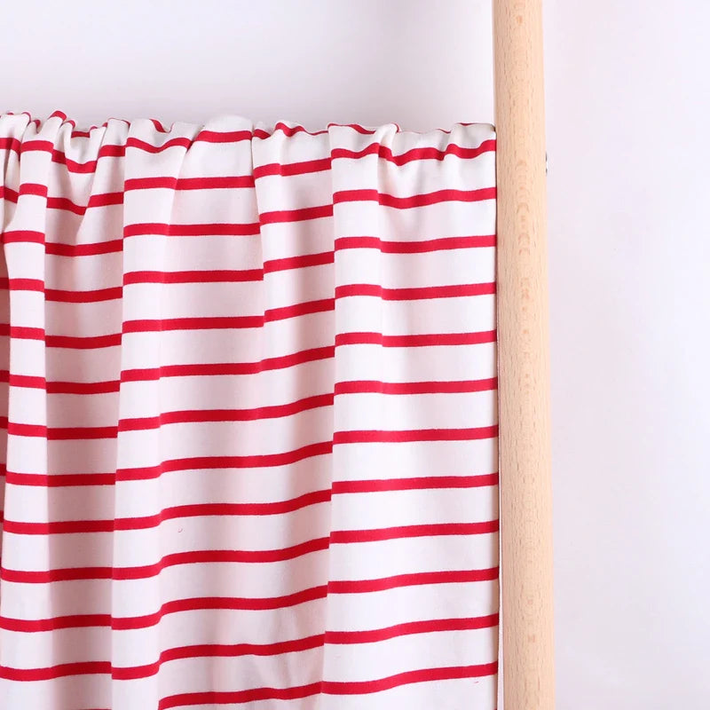 Un rideau blanc à rayures rouges horizontales en Tissu French Terry extensible rayé coloré de Longan Craft France est suspendu à une tringle en bois clair contre un mur blanc uni.