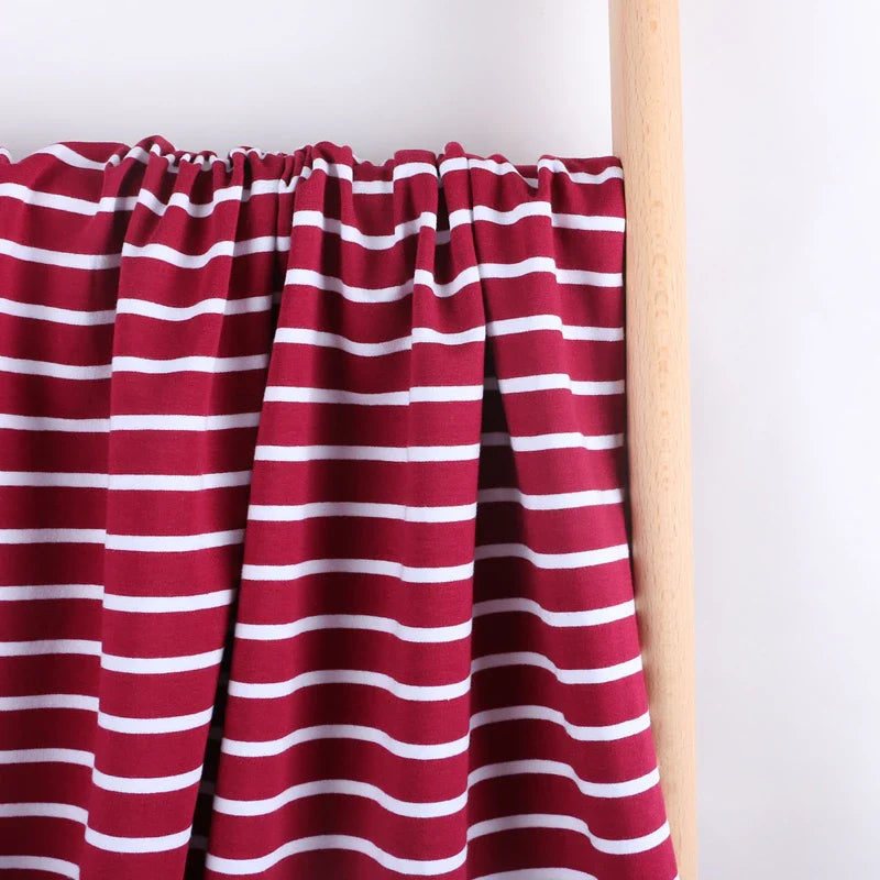 Le Tissu French Terry extensible rayé coloré de Longan Craft France, en coton tricoté à rayures blanches sur fond bordeaux, est drapé sur un poteau en bois clair devant un mur blanc.