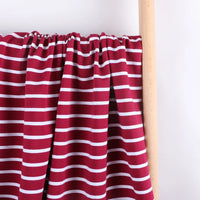 Le Tissu French Terry extensible rayé coloré de Longan Craft France, en coton tricoté à rayures blanches sur fond bordeaux, est drapé sur un poteau en bois clair devant un mur blanc.
