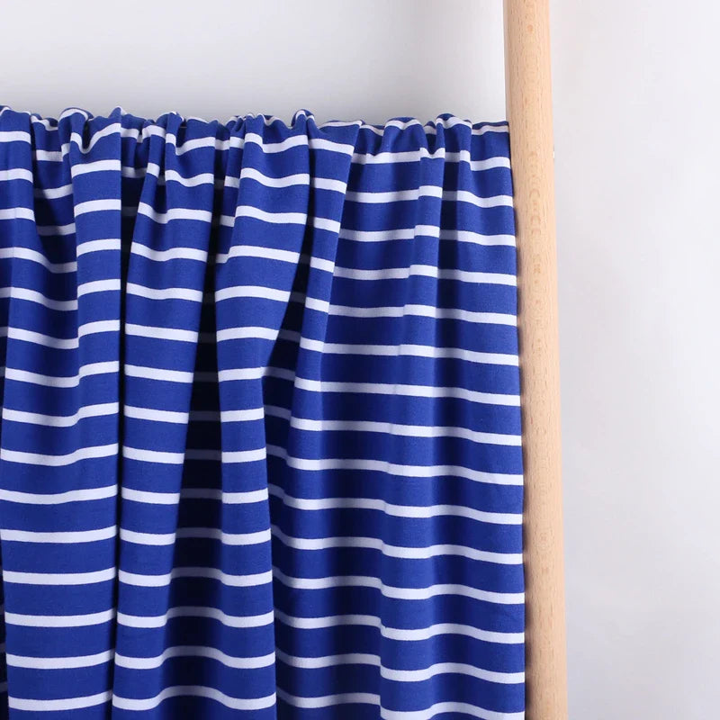 Gros plan sur le Tissu French Terry extensible rayé coloré au mètre de Longan Craft France, qui présente un tricot de coton bleu avec des rayures blanches horizontales, drapé sur une tige en bois sur un fond blanc uni.