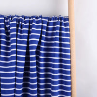 Gros plan sur le Tissu French Terry extensible rayé coloré au mètre de Longan Craft France, qui présente un tricot de coton bleu avec des rayures blanches horizontales, drapé sur une tige en bois sur un fond blanc uni.