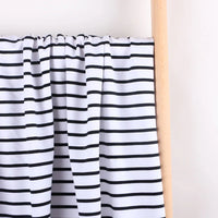 Le Longan Craft France Tissu French Terry extensible rayé coloré au mètre, un tissu en coton tricoté idéal pour sweatshirts, est suspendu sur une barre en bois devant un fond clair.