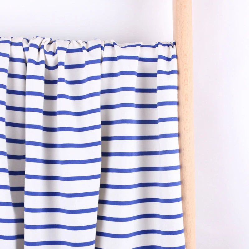 Le "Tissu French Terry extensible rayé coloré" de Longan Craft France, un tricot de coton blanc à rayures bleues, est drapé sur une tige de bois clair sur un fond blanc uni.