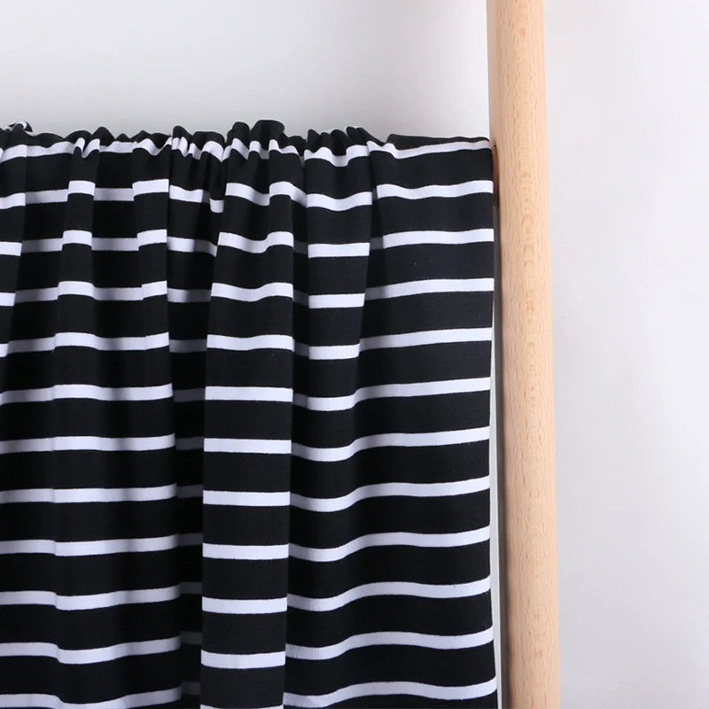 Le French Terry extensible rayé coloré au mètre de Longan Craft France, un tricot de coton idéal pour les sweat-shirts, est présenté en noir avec des rayures horizontales blanches, drapé sur une tige en bois clair sur un fond blanc uni.