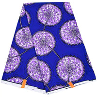 Un morceau plié de tissu wax africain Ankara coloré de Longan Craft France présente un fond bleu vif avec de grandes fleurs de pissenlit stylisées, des tiges noires, des accents blancs et deux étiquettes orange visibles sur le bord.