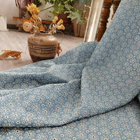 Un morceau de Tissu jean brodé à œillets en coton bleu clair de Longan Craft France, orné d'un motif floral ajouré, est posé sur un sol en bois près d'un vase marron à motifs avec des fleurs séchées. Des plantes vertes apportent une touche de fraîcheur.
