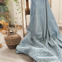 Un grand morceau de Tissu jean brodé à œillets en coton bleu clair 100 % by Longan Craft France avec un motif floral est drapé sur un sol en bois clair à côté d'un vase marron avec des fleurs séchées, un miroir et des plantes vertes en arrière-plan.