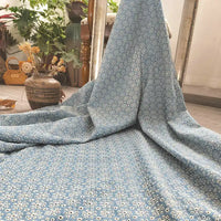 Un tissu bleu clair 100 % coton, Tissu jean brodé floral ajouré de Longan Craft France, est drapé sur une chaise en bois et étalé sur le sol, avec un vase en céramique contenant des fleurs séchées et un sac en osier à l'arrière-plan.