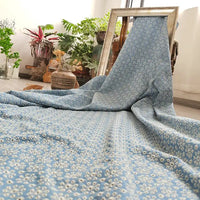 Un tissu denim bleu clair 100 % coton, "Tissu jean brodé floral ajouré" de Longan Craft France, est drapé dans un cadre rectangulaire sur le sol. Des plantes en pot et des objets de décoration apparaissent dans l'arrière-plan intérieur éclairé par le soleil.