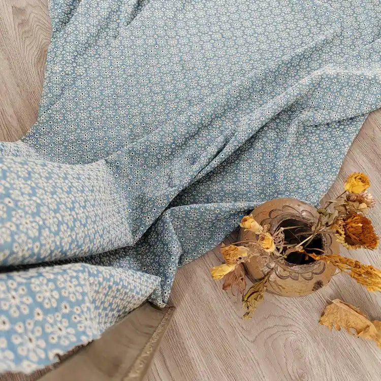 Un tissu denim brodé à œillets en coton bleu clair, "Tissu jean brodé à œillets en coton" de Longan Craft France, est étalé sur un plancher en bois à côté d'un vase de fleurs séchées. La vue de dessus met en valeur son délicat motif ajouré et son aspect rustique.