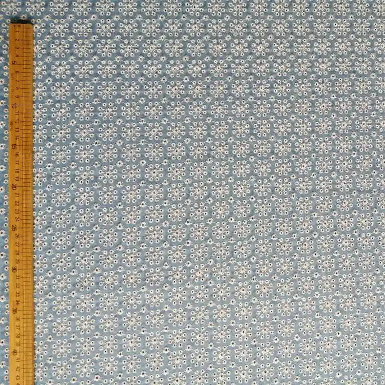 Une règle en bois se tient verticalement le long du bord gauche d'un tissu denim bleu clair 100 % coton avec des œillets floraux brodés, Tissu jean brodé à œillets par Longan Craft France.