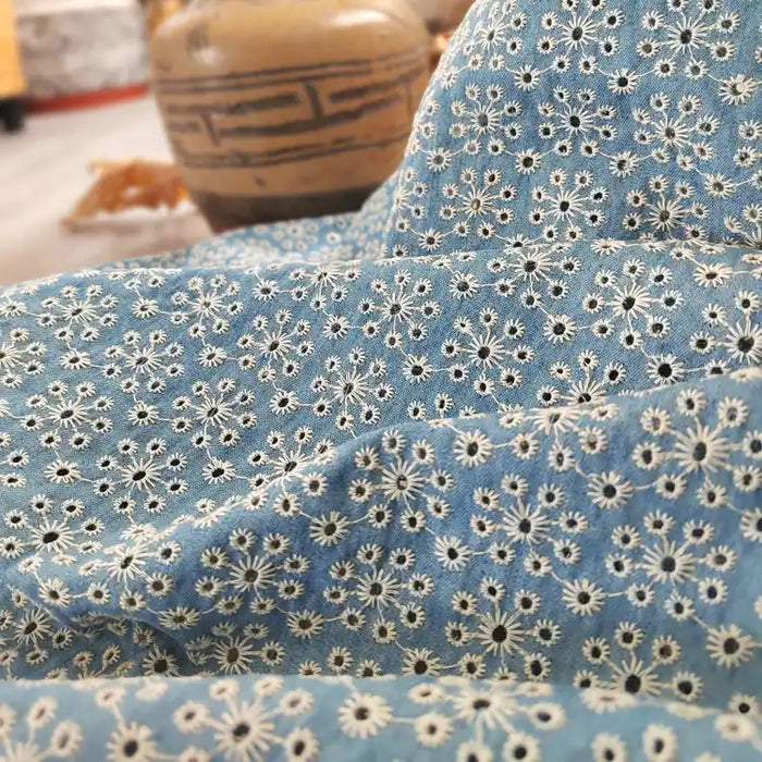 Gros plan sur le tissu jean brodé à œillets en coton bleu clair de Longan Craft France, un tissu denim léger 100 % coton, orné de motifs floraux ajourés ; le fond montre un pot en céramique flou et un objet marron indistinct.