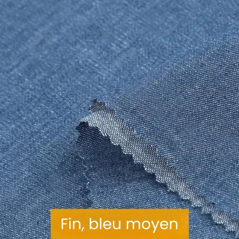 Gros plan sur un tissu denim coton texturé bleu clair avec un bord coupé en zigzag. En bas, une étiquette jaune indique "Fin, bleu moyen". Produit : Tissu denim coton au mètre de Longan Craft France - denim disponible en différents poids et couleurs.
