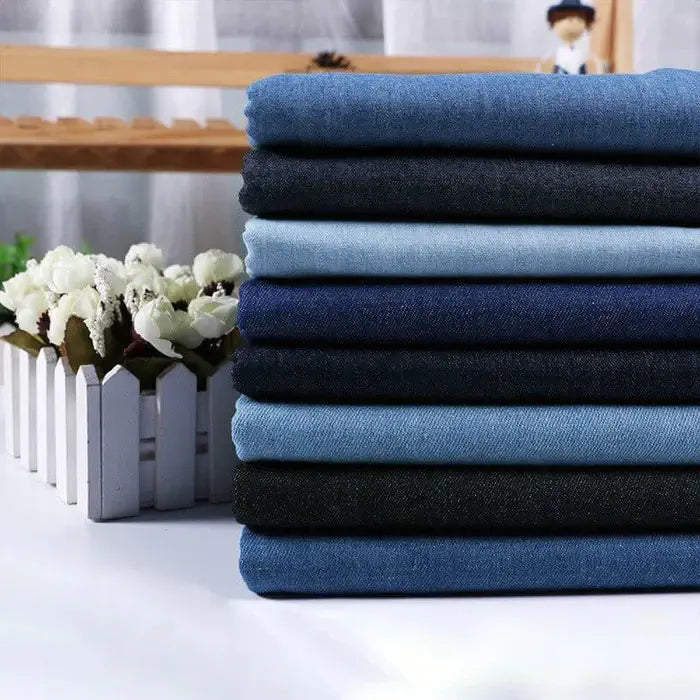 Une pile de Tissu denim coton au mètre de Longan Craft France dans différentes nuances de bleu et de noir est présentée sur une surface blanche, avec un bac à fleurs blanc en arrière-plan. Le denim est disponible en différents grammages.