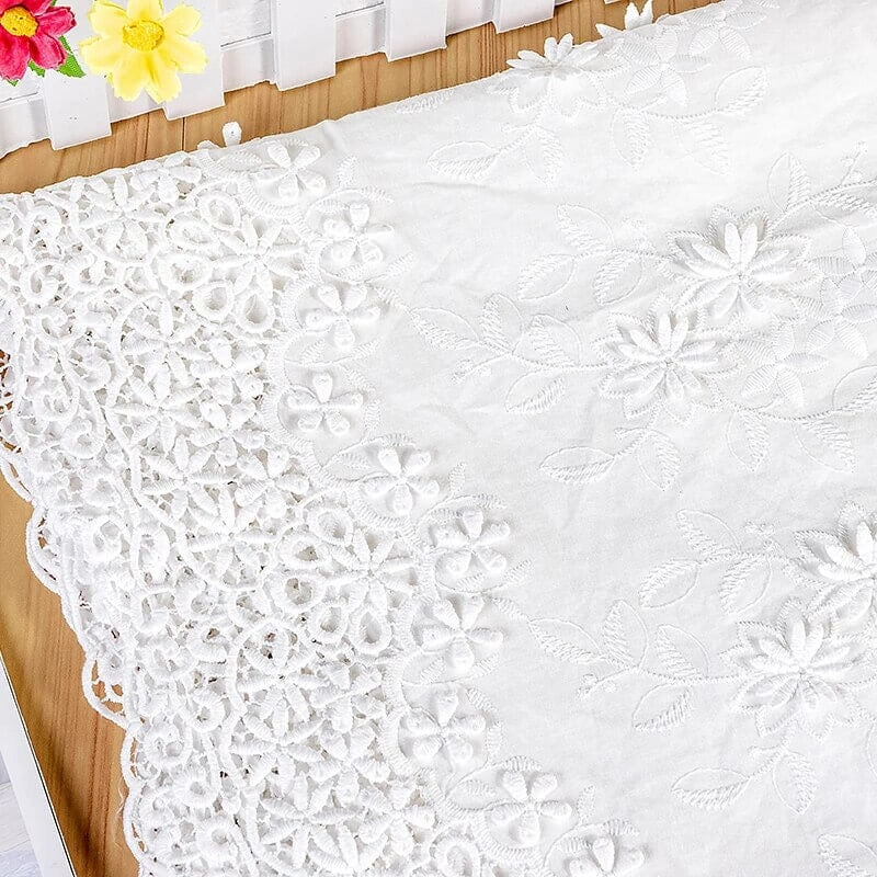 Gros plan sur l'œillet au mètre en tissu en coton ajouré 3D brodé de Longan Craft France, montrant ses motifs floraux complexes, présenté sur du bois près d'une clôture blanche avec des fleurs artificielles jaunes et roses. Convient aux vêtements et aux décors raffinés.