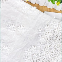 Gros plan sur le tissu en coton ajouré 3D brodé œillet au mètre de Longan Craft France, idéal pour les vêtements raffinés et les textiles décoratifs, qui présente des broderies et des dentelles complexes sur une surface en bois près d'une clôture blanche et de feuilles vertes.