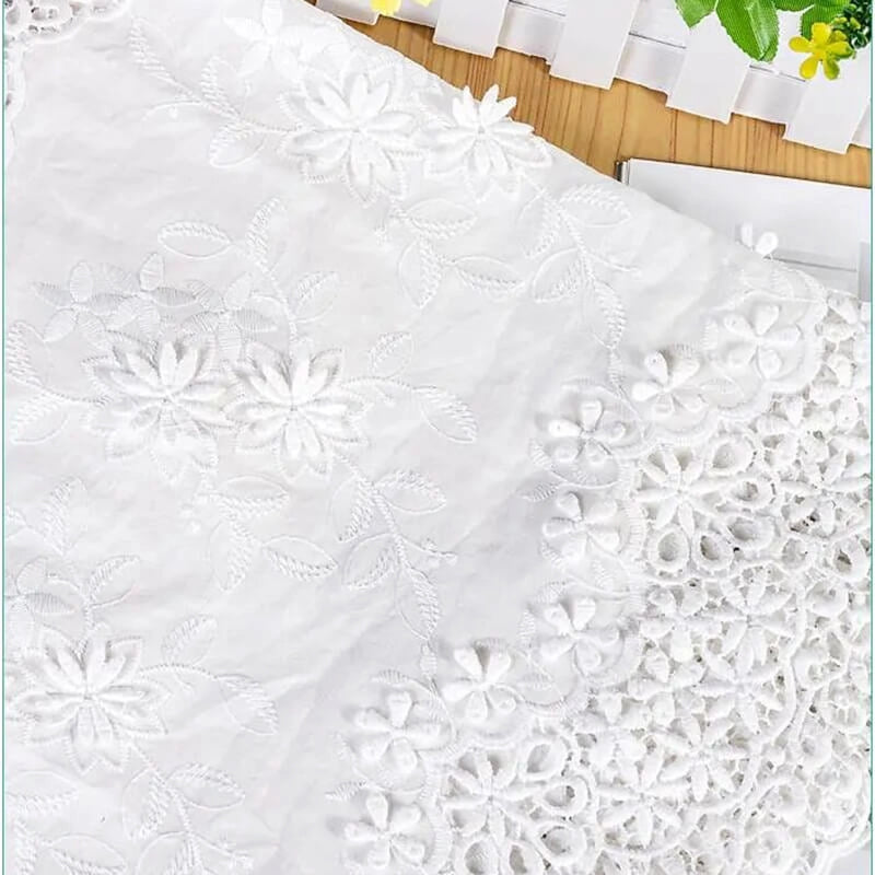 Gros plan sur le tissu en coton ajouré 3D brodé œillet au mètre de Longan Craft France, idéal pour les vêtements raffinés et les textiles décoratifs, qui présente des broderies et des dentelles complexes sur une surface en bois près d'une clôture blanche et de feuilles vertes.