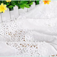 Un gros plan du tissu en coton ajouré 3D brodé œillet au mètre de Longan Craft France est drapé par une clôture blanche, avec des feuilles vertes et des fleurs jaunes en arrière-plan. Parfait pour les vêtements raffinés et les textiles décoratifs.