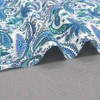 A léger et doux tissu popeline de coton motif paisley bleu et vert de Longan Craft France est posé sur une surface grise, révélant des ondulations subtiles. Parfait pour vêtements, chemises et accessoires chics.