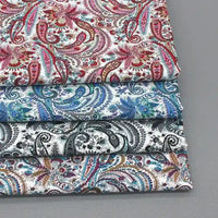 Trois pièces pliées de Tissu popeline de coton motif paisley au mètre de Longan Craft France, dans des tons rouges, bleus et noirs, avec des motifs floraux paisley complexes, sont empilées sur une surface grise. Idéal pour les vêtements et accessoires chics.