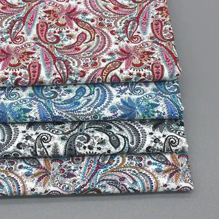 Trois pièces pliées de Tissu popeline de coton motif paisley au mètre de Longan Craft France, dans des tons rouges, bleus et noirs, avec des motifs floraux paisley complexes, sont empilées sur une surface grise. Idéal pour les vêtements et accessoires chics.