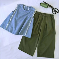 Un haut bleu sans manches avec des bordures blanches est stylisé avec un pantalon vert olive en utilisant le Patron de couture pantalon capri à jambes larges PDF de Longan Craft France. Des ciseaux noirs et du fil vert complètent cette création de mode pour débutants.