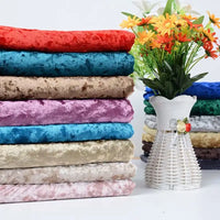 Une sélection de Tissu en velours froissé au mètre de Longan Craft France, un tissu de velours doux de 63 pouces de large pour l'ameublement et le bricolage, est empilée à côté d'un vase en osier blanc avec des fleurs artificielles jaunes et orange sur un fond blanc uni.