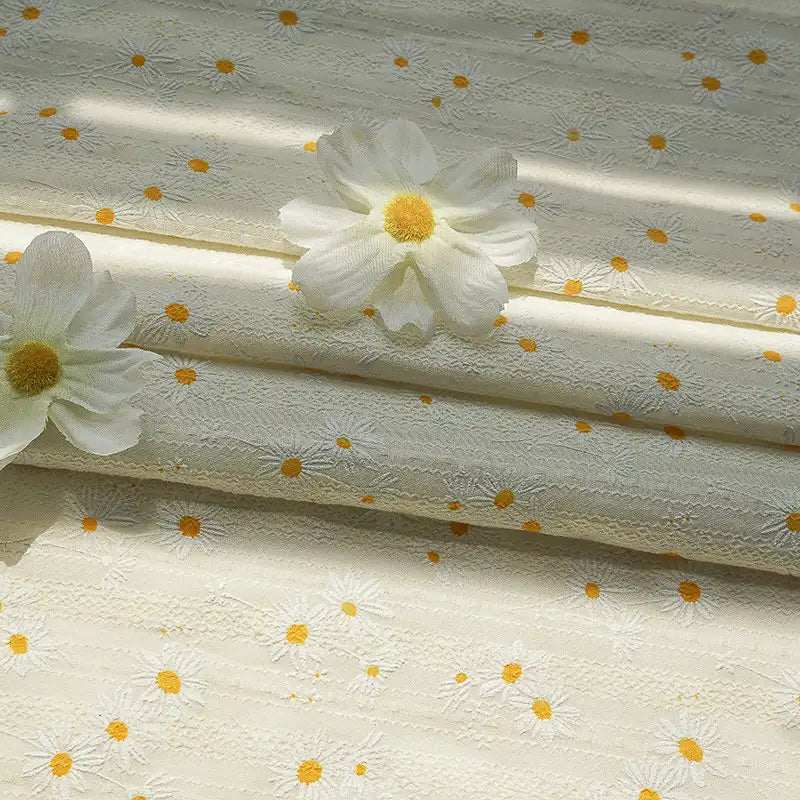 La photo éclairée uniformément montre le tissu dobby imprimé marguerites au mètre de Longan Craft France - un tissu de coton texturé de couleur crème avec un motif floral de marguerites. Deux marguerites artificielles blanches à centre jaune reposent sur le tissu.