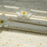La photo éclairée uniformément montre le tissu dobby imprimé marguerites au mètre de Longan Craft France - un tissu de coton texturé de couleur crème avec un motif floral de marguerites. Deux marguerites artificielles blanches à centre jaune reposent sur le tissu.