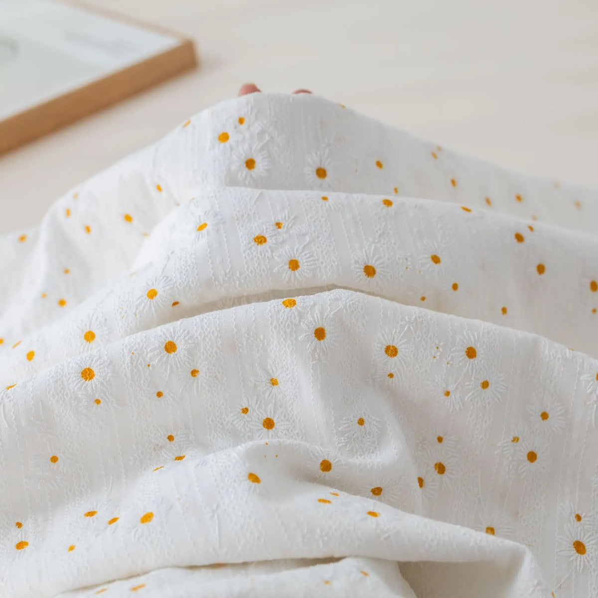 Le Tissu dobby imprimé marguerites au mètre de Longan Craft France est un tissu de coton blanc texturé avec de petites fleurs jaunes, parfait pour réaliser des vêtements confortables ou des projets de décoration. Un livre apparaît partiellement derrière le tissu.