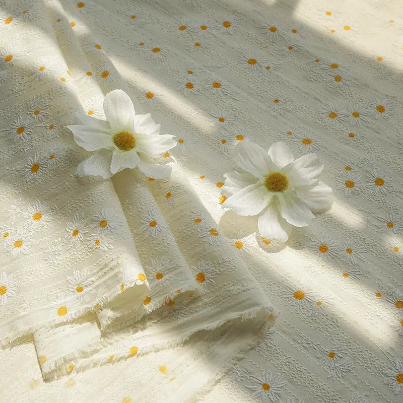 Le tissu dobby imprimé marguerites de Longan Craft France, en coton texturé, présente de petites fleurs jaunes et blanches. Idéal pour vêtements d'été, il est confortable et vendu au mètre : parfait pour vos créations estivales.