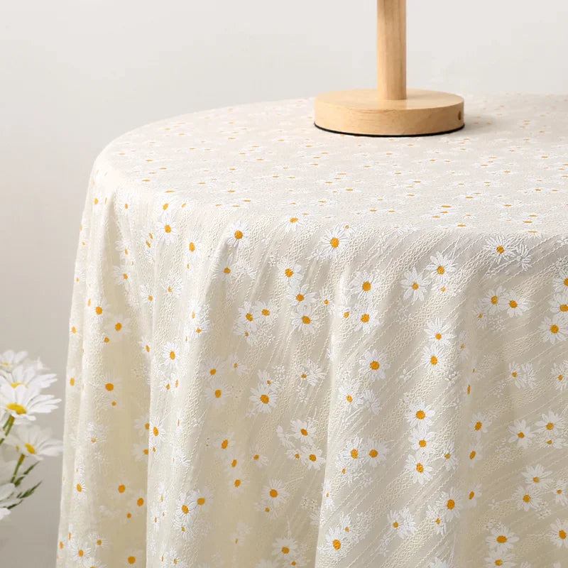 Une table ronde est drapée de Tissu dobby imprimé marguerites de Longan Craft France, un tissu de coton floral crème avec des marguerites blanches et jaunes. Un support en bois est posé sur la table et un vase avec des marguerites apparaît à gauche.