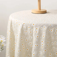 Une table ronde est drapée de Tissu dobby imprimé marguerites de Longan Craft France, un tissu de coton floral crème avec des marguerites blanches et jaunes. Un support en bois est posé sur la table et un vase avec des marguerites apparaît à gauche.