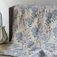 Le Tissu brocart pissenlit brodé de Longan Craft France, un jacquard métallique de couleur crème avec des broderies florales bleues et vertes et des accents dorés, est drapé sur une surface brune à côté d'un vase géométrique blanc et d'une bouteille en verre transparent.