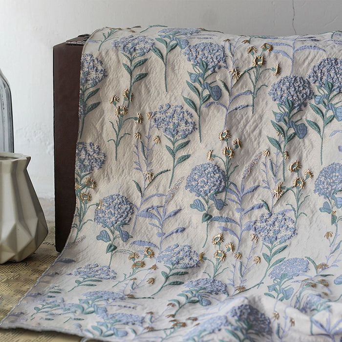 Le Tissu brocart pissenlit brodé de Longan Craft France, un jacquard métallique de couleur crème avec des broderies florales bleues et vertes et des accents dorés, est drapé sur une surface brune à côté d'un vase géométrique blanc et d'une bouteille en verre transparent.