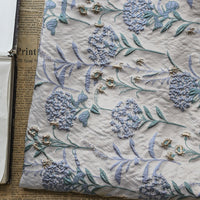 Un gros plan du Tissu brocart pissenlit brodé de Longan Craft, un jacquard d'ameublement floral vintage avec des fils métalliques, montre des détails brodés bleus et verts en relief sur le tissu texturé posé sur du papier journal.