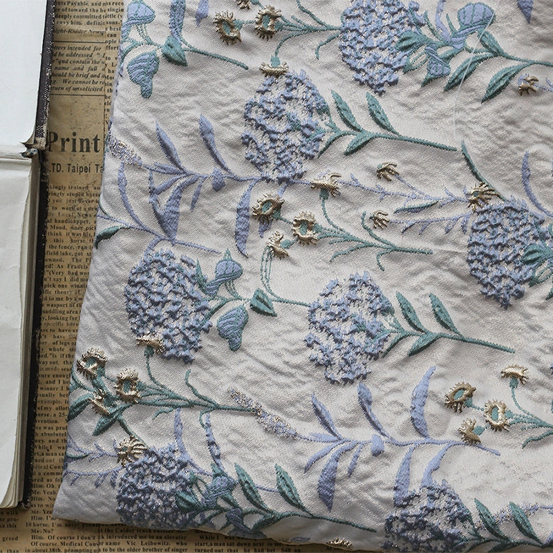 Un gros plan du Tissu brocart pissenlit brodé de Longan Craft, un jacquard d'ameublement floral vintage avec des fils métalliques, montre des détails brodés bleus et verts en relief sur le tissu texturé posé sur du papier journal.