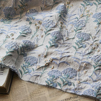 Un morceau de Tissu brocart pissenlit brodé de Longan Craft France, un tissu d'ameublement jacquard métallique floral vintage, est drapé sur des livres empilés et de vieilles pages de journaux.