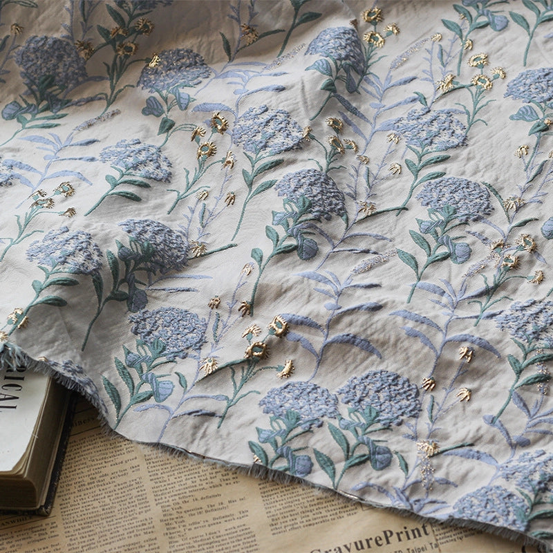 Un morceau de Tissu brocart pissenlit brodé de Longan Craft France, un tissu d'ameublement jacquard métallique floral vintage, est drapé sur des livres empilés et de vieilles pages de journaux.