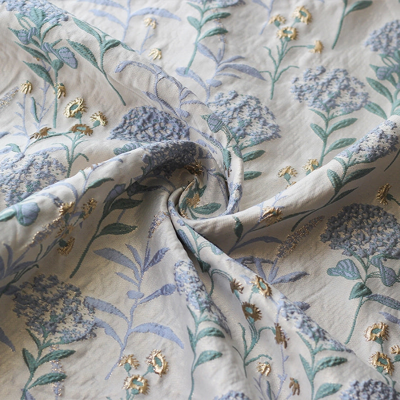 Un gros plan du Tissu brocart pissenlit brodé de Longan Craft France montre son motif jacquard floral dans les tons bleu, vert et or. Doucement torsadé au centre, le tissu révèle des fleurs et des feuilles brodées avec des détails de damas métallique.