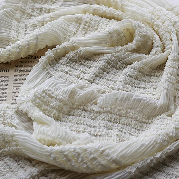 Gros plan sur le tissu mousseline plissé extensible blanc de Longan Craft France, doux et diaphane avec une texture plissée, drapé sur un journal. Idéal pour les robes, ce tissu léger est vendu au mètre en blanc, abricot, rose ou noir.