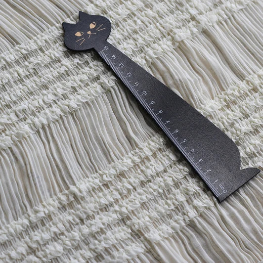 Une règle noire en forme de chat repose sur un tissu mousseline stretch plissé blanc (Longan Craft France), idéal pour les robes plissées, présenté ici dans l'option de couleur uni blanc.