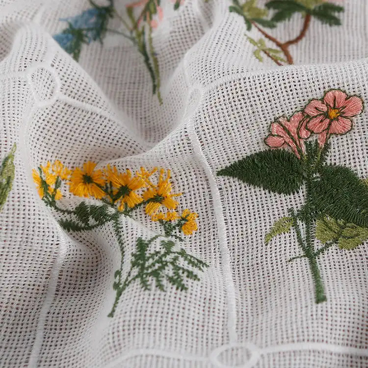 Gros plan sur le Tissu coton brodé de motifs floraux de Longan Craft France, un tissu de coton à carreaux doux pour l'habillement, présentant des fleurs brodées colorées - des fleurs jaunes et roses avec des tiges vertes sur une surface texturée.