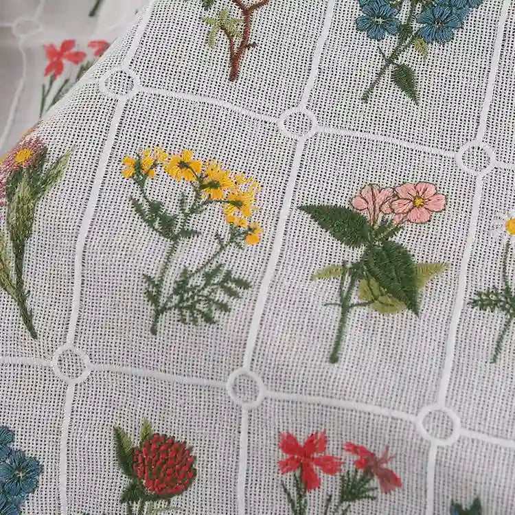 Gros plan sur le "Tissu coton brodé de motifs floraux" de Longan Craft France : tissu de coton blanc à motif quadrillé, avec des fleurs colorées brodées dans chaque carré. Matière douce, parfaite pour la confection de vêtements.