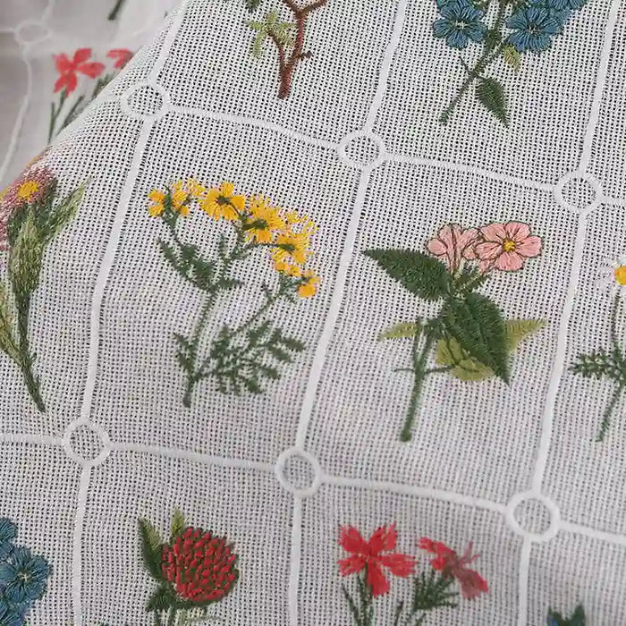 Gros plan sur le "Tissu coton brodé de motifs floraux" de Longan Craft France : tissu de coton blanc à motif quadrillé, avec des fleurs colorées brodées dans chaque carré. Matière douce, parfaite pour la confection de vêtements.
