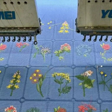 Deux machines à broder cousent des motifs floraux colorés sur un Tissu coton brodé de motifs floraux à carreaux de Longan Craft France, un tissu de coton doux idéal pour l'habillement.